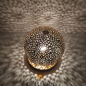 K&ouml;nnte beinhalten: Eine runde, goldfarbene marokkanische Laterne mit filigranen Ausschnitten. Die Laterne ist beleuchtet und strahlt ein warmes Licht aus. Der Text "ATLAS LIGHTS MOROCCAN LIGHTING" ist unterhalb der Laterne sichtbar.