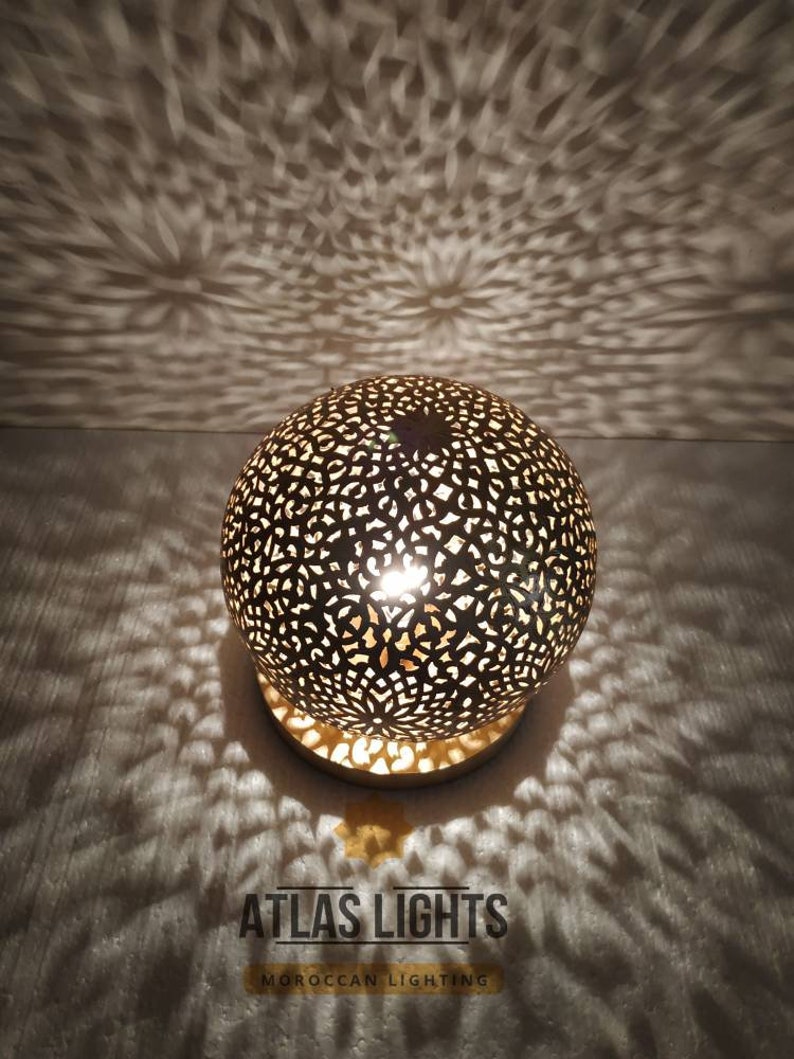 K&ouml;nnte beinhalten: Eine runde, goldfarbene marokkanische Laterne mit filigranen Ausschnitten. Die Laterne ist beleuchtet und strahlt ein warmes Licht aus. Der Text "ATLAS LIGHTS MOROCCAN LIGHTING" ist unterhalb der Laterne sichtbar.