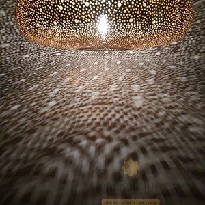 May include: A large, round, gold-colored Moroccan pendant light fixture with a decorative, intricate, cut-out pattern. The light fixture is hanging from the ceiling and casting a pattern of light on the floor below. The light fixture is made of metal and has a warm, inviting glow.