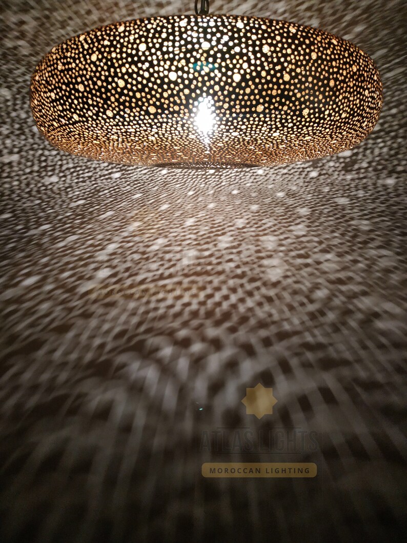 May include: A large, round, gold-colored Moroccan pendant light fixture with a decorative, intricate, cut-out pattern. The light fixture is hanging from the ceiling and casting a pattern of light on the floor below. The light fixture is made of metal and has a warm, inviting glow.