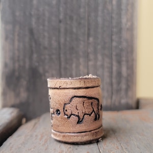 Könnte beinhalten: Eine zylindrische Perle aus beigem Leder mit einer handgefertigten Bison-Silhouette und einem kleinen sternförmigen Stempeldesign. Das Leder hat ein natürliches, hellbraunes Finish mit dunklen, eingebrannten Details, die die Tierform betonen.