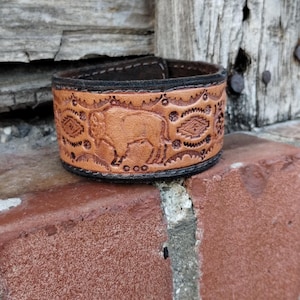 May include: A brown leather bracelet with a tooled design of a buffalo and geometric patterns. The bracelet has a black stitching detail.