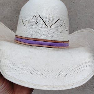 K&ouml;nnte beinhalten: Ein wei&szlig;er Strohhut im Cowboy-Stil mit einem purpurfarbenen und braunen Lederband.