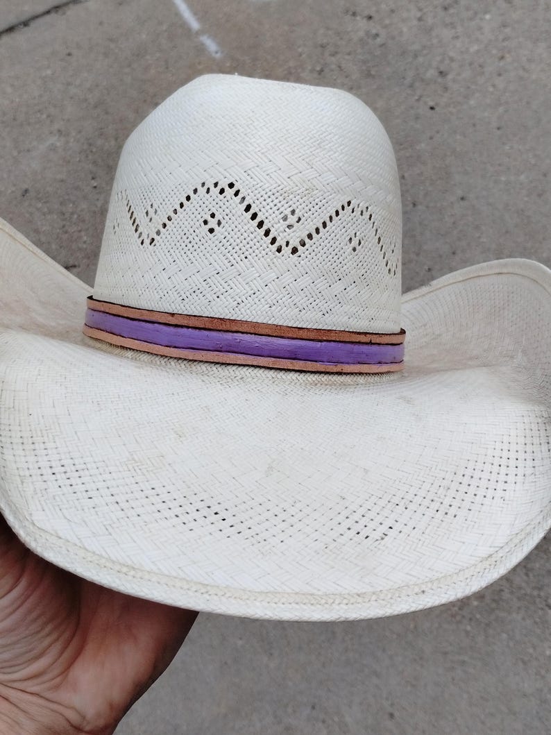 K&ouml;nnte beinhalten: Ein wei&szlig;er Strohhut im Cowboy-Stil mit einem purpurfarbenen und braunen Lederband.