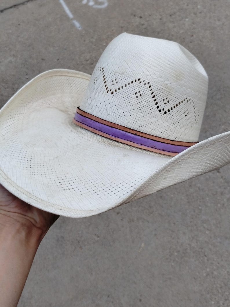 K&ouml;nnte beinhalten: Wei&szlig;er Strohhut im Cowboy-Stil mit einem lila und braunen Band. Der Hut hat ein gewebtes Design und eine breite Krempe.