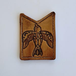 May include: A rectangular, light brown leather item with a carved bird design. The bird has outstretched wings and a cross-like pattern down its center. The edges are stitched with white thread.