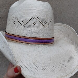 K&ouml;nnte beinhalten: Wei&szlig;er Strohhut im Cowboy-Stil mit einem violetten und braunen Band um die Krone.