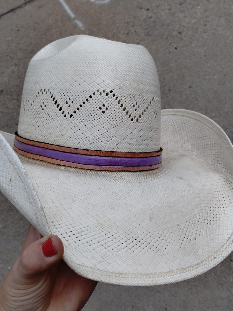 K&ouml;nnte beinhalten: Wei&szlig;er Strohhut im Cowboy-Stil mit einem violetten und braunen Band um die Krone.
