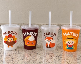 May include: Four clear plastic tumblers with lids and straws, each featuring a unique autumn-themed graphic and a name. The designs include a turkey labelled Aurora, a pumpkin pie slice labelled Maeve, a fox labelled Jason, and a scarecrow labelled Mateo.