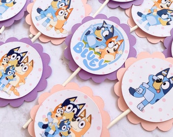 May include: A set of 10 Bluey cupcake toppers featuring different images of the cartoon characters Bluey and Bingo. The toppers are printed on white cardstock with a pink and purple scalloped border. The toppers are perfect for a Bluey themed birthday party.