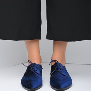 May include: A pair of blue velvet lace-up shoes with pointed toes. The shoes have a black lace and are worn by a person with bare legs.
