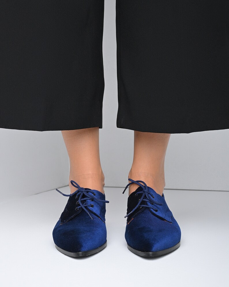 May include: A pair of blue velvet lace-up shoes with pointed toes. The shoes have a black lace and are worn by a person with bare legs.