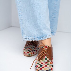May include: Multi-colored checkered pattern shoes with brown leather accents and laces. The shoes have a classic oxford style and are worn with light blue jeans.