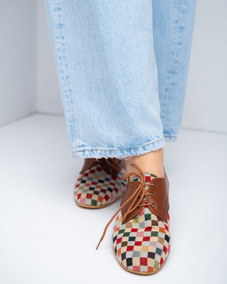 May include: Multi-colored checkered pattern shoes with brown leather accents and laces. The shoes have a classic oxford style and are worn with light blue jeans.