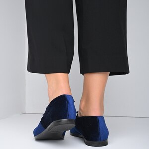 May include: A pair of blue velvet lace-up shoes with black soles. The shoes are worn by a person wearing black pants.