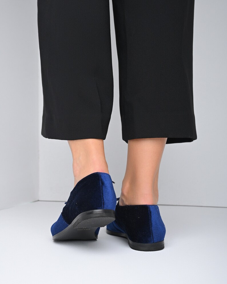 May include: A pair of blue velvet lace-up shoes with black soles. The shoes are worn by a person wearing black pants.