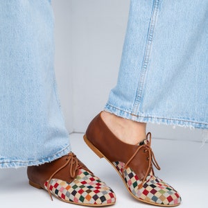 May include: Brown leather lace-up shoes with a colorful, checkered fabric design on the toe and vamp. The shoes have a low heel and are shown with light blue jeans. The shoes are a unique and stylish footwear option.
