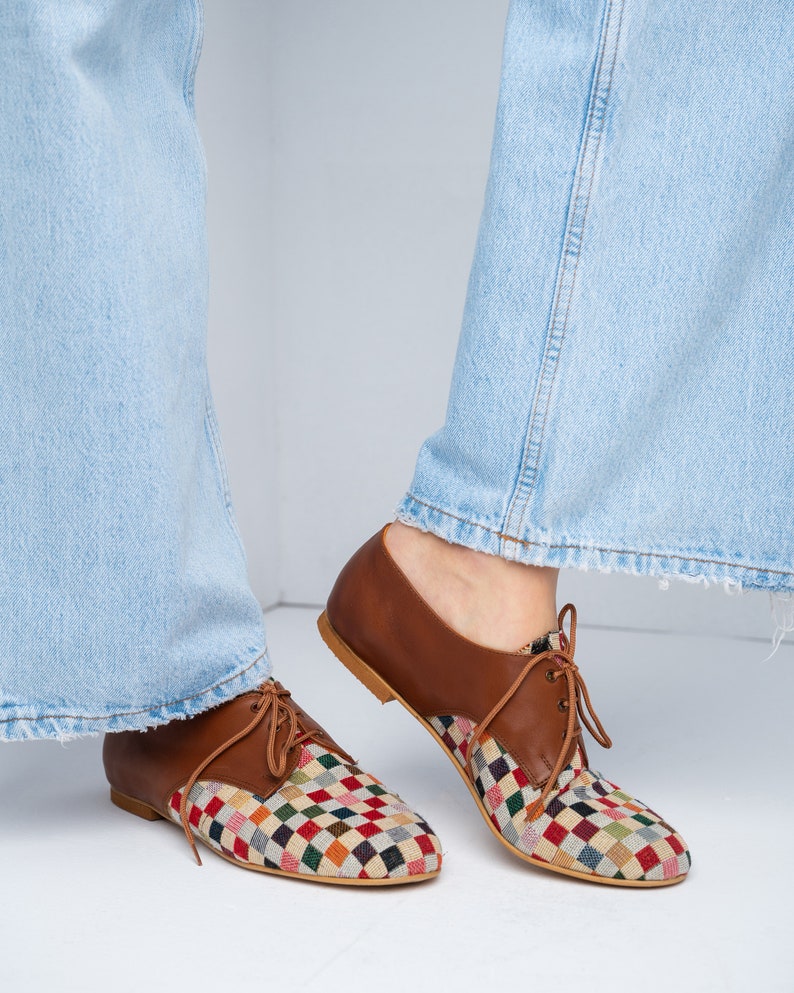 May include: Brown leather lace-up shoes with a colorful, checkered fabric design on the toe and vamp. The shoes have a low heel and are shown with light blue jeans. The shoes are a unique and stylish footwear option.