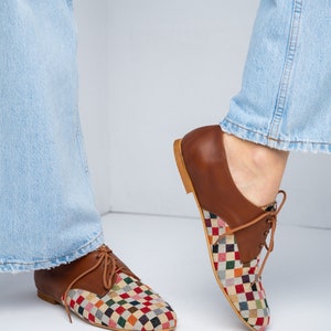 May include: Brown leather oxford shoes with a colorful checkered fabric design on the toe and vamp. The shoes have a classic lace-up closure and a light brown sole. The shoes are shown with light blue jeans.