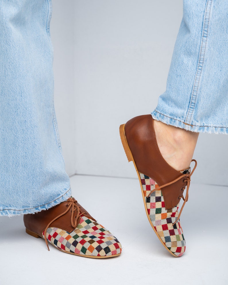 May include: Brown leather oxford shoes with a colorful checkered fabric design on the toe and vamp. The shoes have a classic lace-up closure and a light brown sole. The shoes are shown with light blue jeans.