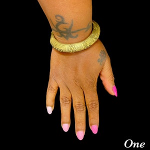May include: A gold bangle bracelet with an intricate design. The bracelet is worn on a person's wrist with pink nail polish.