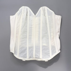May include: A white corset with a scalloped neckline and vertical boning. The corset is made of a sheer fabric with opaque vertical panels and delicate lace detailing at the top. The garment is designed to cinch the waist and provide support.