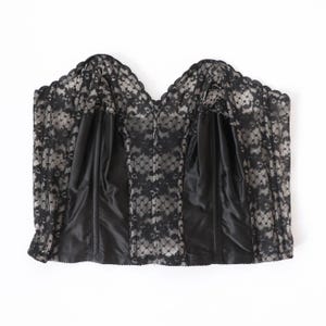 May include: A strapless corset top featuring black floral lace overlays on a solid black satin base. The garment has a sweetheart neckline with a scalloped edge, vertical boning channels, and a structured bodice design with a straight hemline.