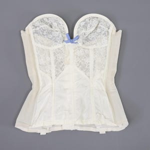 May include: A white strapless corset with a blue and white bow at the centre. The corset is made of lace and has a smooth, satin-like finish.