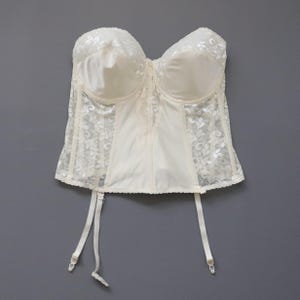 May include: Ivory-colored corset with lace detailing. The bust features padded cups, and the garment includes attached garter straps. The corset is made of a satin-like material and lace, with a scalloped edge.