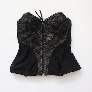 May include: Black corset featuring a lace overlay on the bust and central panel. It has a front zip closure and a ribbon tie at the top. The edges are trimmed with delicate lace.