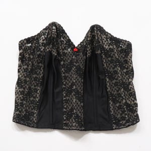 May include: A strapless corset top featuring black floral lace overlays on a beige base. The garment has vertical black fabric panels, a scalloped neckline, and a small red bow detail centered at the top edge of the bodice for a structured look.