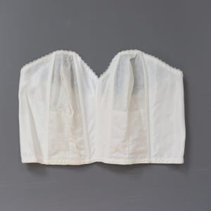 May include: A white strapless corset with a scalloped edge. The corset has vertical seams and a structured design. The garment is made of a smooth, opaque fabric and is displayed against a gray background.