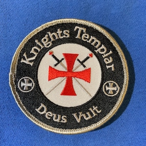 May include: A black and white embroidered patch with a red cross and two swords. The text "Knights Templar" is written in a circle around the cross and swords. The text "Deus Vult" is written below the cross and swords.