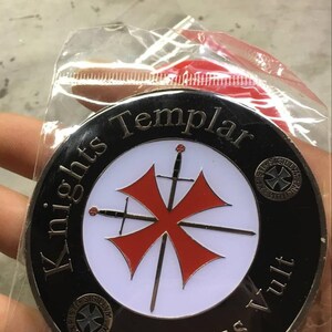 Puede incluir: Una moneda redonda, negra y plateada con las palabras "Knights Templar" y "Deus Vult". La moneda tiene un centro blanco con una cruz roja y dos espadas. La moneda est&aacute; en una bolsa de pl&aacute;stico transparente.