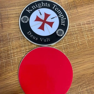 Puede incluir: Un posavasos redondo con base roja y una parte superior con las palabras "Knights Templar" alrededor de un dise&ntilde;o de cruz roja. Tambi&eacute;n est&aacute;n presentes las palabras "Deus Vult". El posavasos tiene un borde negro y est&aacute; sobre una superficie de madera.
