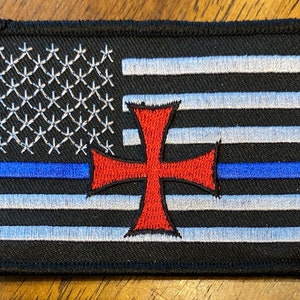 May include: A black and white American flag with a blue stripe and a red cross in the center. The flag is embroidered and has a stitched border.
