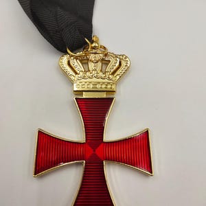 May include: A gold-toned metal cross pendant featuring a vibrant red translucent enamel inlay with a fine ribbed texture. The cross is topped with a detailed gold crown ornament and is attached to a black fabric ribbon for suspension.