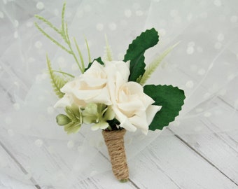 Puede incluir: Un pequeño boutonniere de flores blancas con tres rosas blancas, hojas verdes y una envoltura de cuerda marrón.