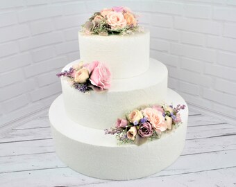 May include: Three-tiered white cake dummy decorated with pink, peach, and purple artificial flowers. The cake is on a white wooden surface.