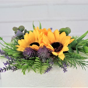 May include: A two-tiered white cake decorated with a sunflower and thistle floral arrangement. The arrangement includes yellow sunflowers, purple thistles, and green foliage.