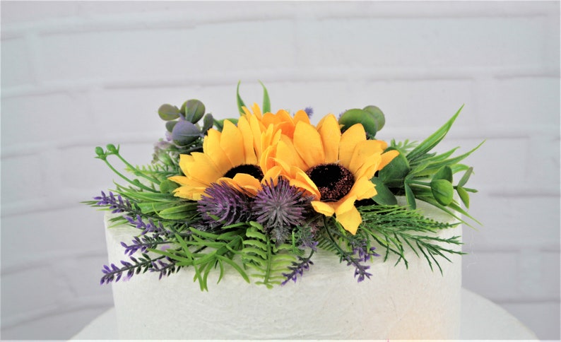 May include: A two-tiered white cake decorated with a sunflower and thistle floral arrangement. The arrangement includes yellow sunflowers, purple thistles, and green foliage.