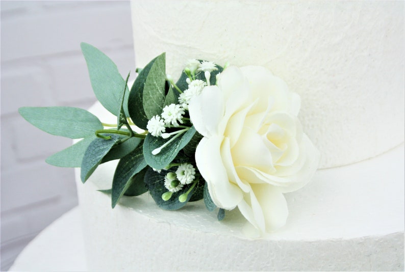 May include: A white cake topper featuring a single white rose, small white flowers, and green leaves.
