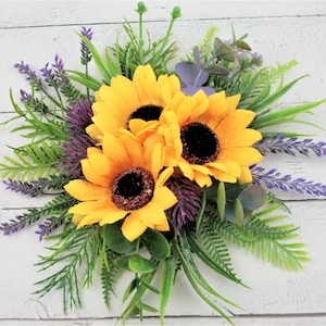 May include: A bouquet of three yellow sunflowers with dark centers, surrounded by green leaves and purple lavender sprigs.