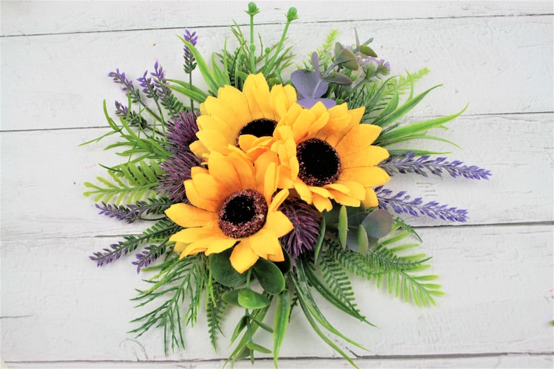 May include: A bouquet of three yellow sunflowers with dark centers, surrounded by green leaves and purple lavender sprigs.