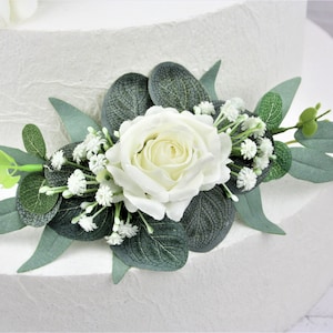 May include: White faux rose with green eucalyptus leaves and small white flowers, a wedding cake topper.