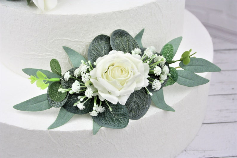 May include: White faux rose with green eucalyptus leaves and small white flowers, a wedding cake topper.