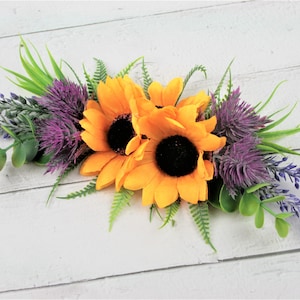 May include: A floral arrangement featuring two yellow sunflowers, purple thistle, and lavender sprigs. The arrangement is on a white wooden background.