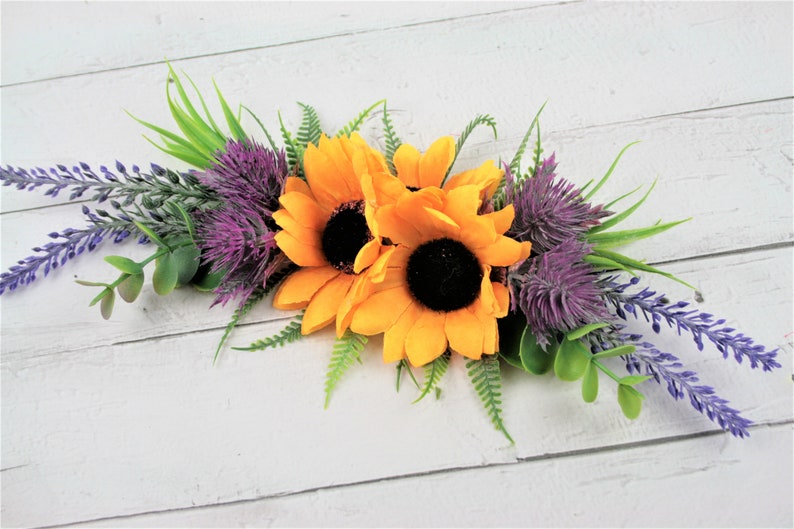 May include: A floral arrangement featuring two yellow sunflowers, purple thistle, and lavender sprigs. The arrangement is on a white wooden background.