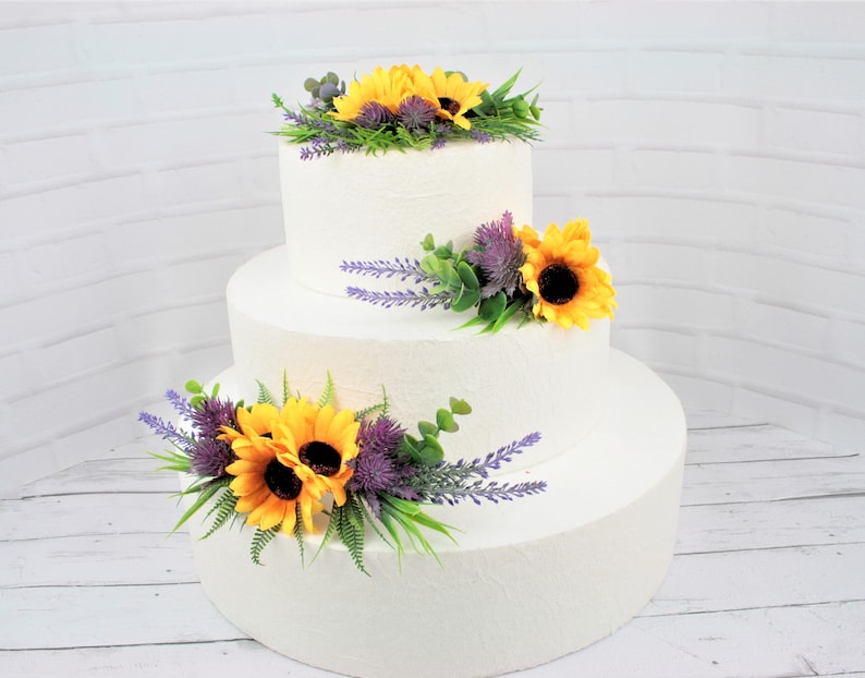 May include: A three-tiered white wedding cake decorated with artificial sunflowers, lavender, and greenery.