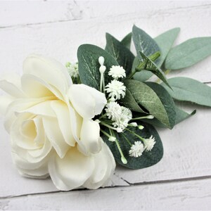 May include: A single white rose with small white flowers and green leaves. The bouquet is arranged on a white wooden surface.